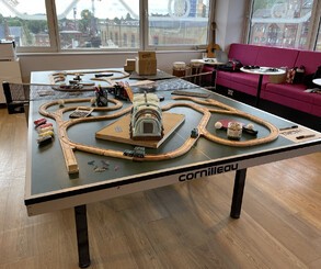 Left: A pool table covered in wooden train tracks and other toys, including a model crane from Tren holding a playing card. Right Top: A ping pong table, also heavily decorated. Right Bottom: A Tren cake!